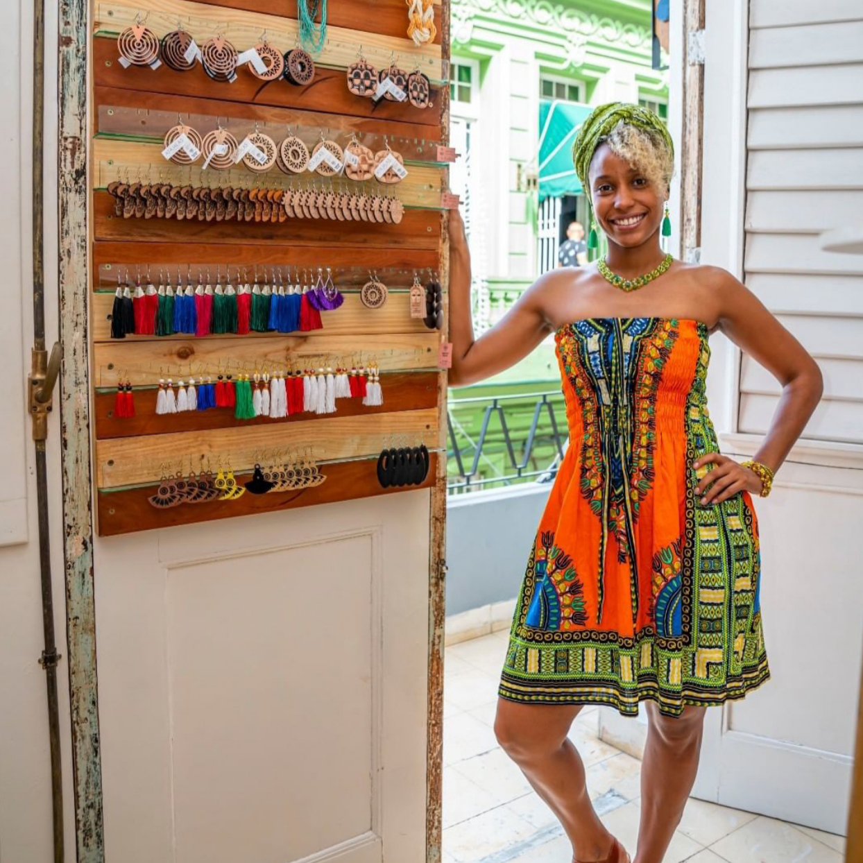 Photo Credit: Beyond Roots woman in colorful African style print dress stands next to wall display of earrings